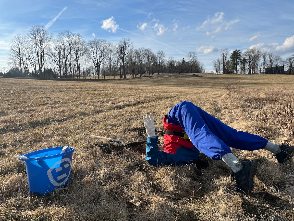 On a vast dried grass field, an artist in blue pants is poking her head in a hole. A shovel and a blue bucket with a smiley face are placed near the artist.